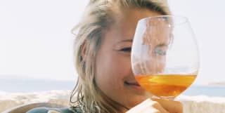 A young woman looks at the camera through the top of a glass of Aperol as she sits in the shade of an olive tree