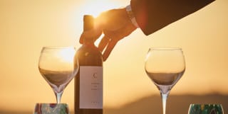 A man places a Castello di Casole private label bottle of red wine on the table, partly silhouetted against the setting sun