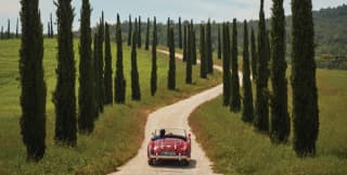 Red vintage soft-top car driving along a winding driveway lined by cypress trees