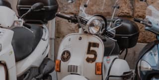Street detail of a row of Vespa motorinos parked against a wall, one with the number 5 emblazoned on its white front panel.