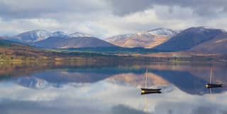 Small fishing boats floating on a mirror-still loch reflecting the snow-capped mountains