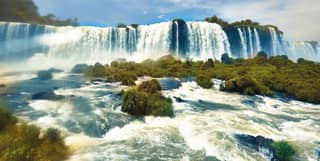 Low angle view from the grassy rock-filled water, as the Iguaçu River tumbles over a wide ledge in impressive cataract falls.