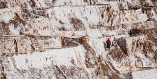 A woman working in the Maras salt mines walks down a steep, narrow path, red clothes contrasting with the white salt ponds.