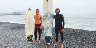 Two surfers standing on a stoney shore holding their surfboards upright