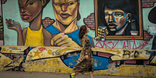 A woman in a bohemian print dress walks past a long Barranco street mural of faces, painted in an angular, arresting style.