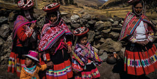 Three women and two children in traditional dress with montera felt hats, stand by a stone wall at a farm in Patacancha.