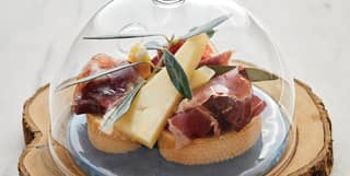 A glass cloche covers a plate of snacks, including slices of baguette topped with Iberico Bellota and Manchego triangles