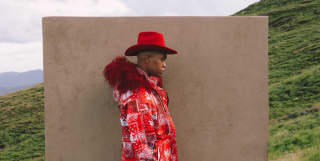 In a hillside magazine fashion shoot, a male model wears a red print jacket with feather trim and Stetson by Thebe Magugu.