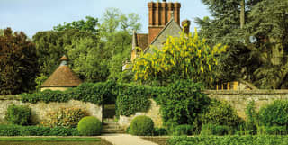 Ivy climbs the garden stone wall. Tall red brick chimney pots and the dovecote's conical roof peer over the tops of tall trees