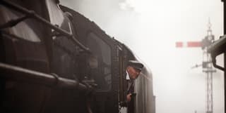 A train driver in a peaked black cap looks out from his cab. The train's body disappearing into the haze of smoke and steam