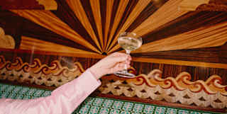 An arm in a pink satin sleeve raises a glass of Champagne up against the stunning marquetry panel behind a green banquette.