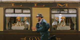 A man, dressed in a blue nineteen-thirties suit and hat runs in a blur on the platform in front of the stationed Gwen car.