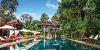 The blue green waters of the hotel swimming pool are surrounded by tall Cambodian trees