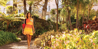 A female guest with dark hair and a short orange and yellow dress walks down a garden path leading away from the hotel.