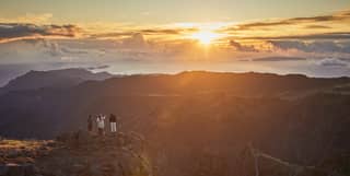 Three guests stand on Madeira's third highest peak, Pico de Arieiro, watching the sun rise over craggy mountains and valleys.