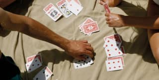 Close-up on a beige beach blanket covered in blue and red-backed playing cards as a group of friends relaxes in the sun.