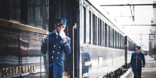 Train guard in a blue uniform blowing a whistle next to train carriages