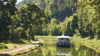 The Fleur de Lys cruises up the serene Ouche river, bordered by big trees and green banks, where a cyclist enjoys the route.