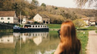 A woman stands on the bank of the canal, taking a photograph of the Fleur de Lys as it cruises by a cluster of white houses.