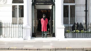 A doorman in a knee-length red coat, black scarf and black bowler, stands between the white pillars of the hotel entrance