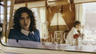 A woman with curly dark hair, in a teal jumper, gazes out of the window by her dining table, seen from outside the train.