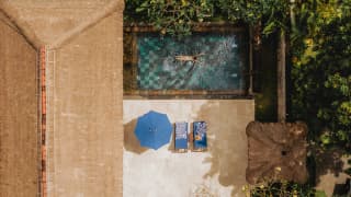 A guest floats in a sapphire pool of a Deluxe Villa, overlooked by a terrace with a parasol and loungers, seen from above.