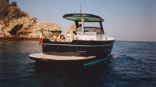 A couple sunbathes on the lounge deck of an Apreamare Gozzo boat as sails in full sun towards the headland, seen from behind.