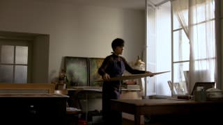 Artist Amélie Chassary, in a dark apron, carries a painting to her workshop table, illuminated by a light-filled window.