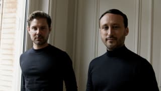 Interior designers Emil Humbert and Christophe Poyet, viewed from the chest up, wearing black, high-neck, long-sleeve shirts.