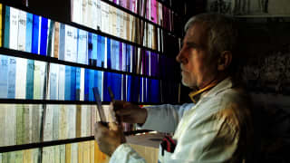 With a grey hair and beard, Bruno Loire regards a backlit wall of stained glass pieces in his Atelier Loire in Chartres.
