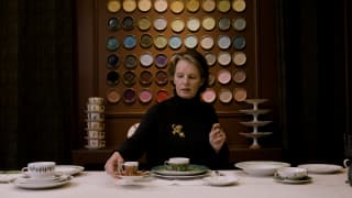 Porcelain painter, Marie Daage, in black, sits behind a table of vintage cups by a wall displaying dishes of varying glazes.