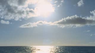 A bleach-white sun in a blue sky with drifting clouds creates a sparkling golden splash on a calm blue ocean.