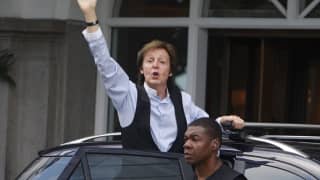 A guard stands by as Sir Paul McCartney in a blue shirt and black waistcoat waves from the open car door outside the hotel.