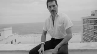 Black and white photo of Freddie Mercury sitting on a roof terrace wall in black pants and white shirt, holding a cigarette.