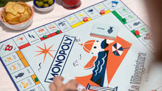 At a table with a red cocktail, olives and crisps, a guest plays Monopoly, the La Villeggiatura edition, seen from above.