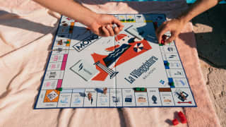 Two guests play Monopoly, La Villeggiatura, sitting either side of the board game on a pink towel by the edge of the pool.