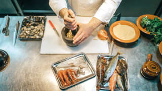 A chef grinds ingredients with a pestle and mortar on a steel kitchen worktop alongside fresh seafood and shellfish