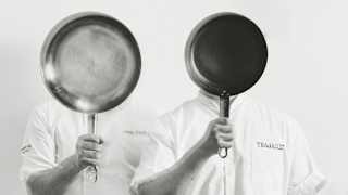 Two people in chef uniforms stand side by side, each holding a frying pan in front of their face, obscuring their features. The image is in black and white against a plain background.
