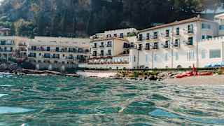 Eye-level view over crystal sea, looking towards Villa Sant'Andrea's white waterfront edifice at the foot of a forested hill.