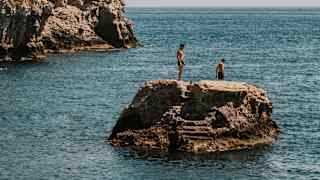 Two figures stand on the sea stack in Mazzarò's small bay contemplating a jump into the water, with the headland seen behind.