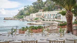 A long table is exquisitely set for lunch on the dining terrace amid palm trees and looking across the Bay at the sweep of hotel