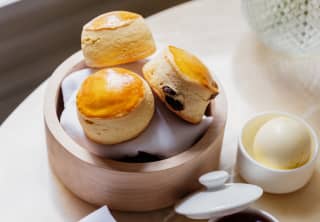 Three fresh, golden-topped fruit scones nestle in a wooden bowl, accompanied by a pots of strawberry jam and clotted cream.