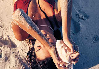 Close-up of a lady in a red swimsuit lying on the sand and holding a conch shell above her head