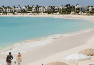 High angle view over the curve and bright blue water of Maundays Bay, where two guests stroll along the soft cream sand.