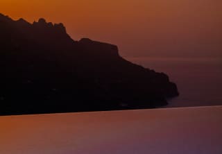 A sunset orange sky melts to red behind the rocky silhouette of the Amalfi coastline, viewed over a sub-lit infinity pool.