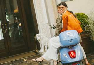 A smiling woman in an orange top and glasses sits side-saddle on the back a blue Vespa motorcycle behind a man in tweed.
