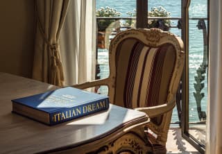 A blue hardback Italian Dream, its title embossed in gold, sits beside an antique chair. Through windows, sun dances on water
