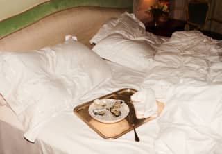 Four fresh oysters lie in a bowl of ice on a golden tray with two forks. The tray sits ready on a recently vacated bed