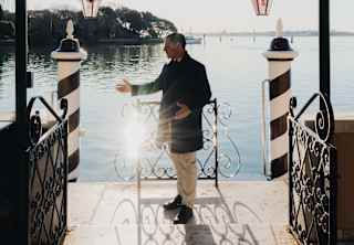 Sun dazzles on the water in front of the hotel, where a staff member in a black jacket stands between the monochrome paline.