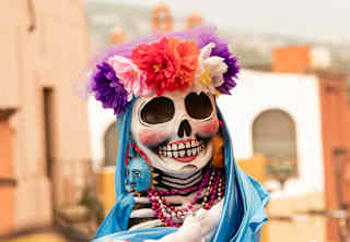 A skull-faced puppet wearing a red, pink and purple floral headband and blue cloak is held above crowds in a street parade.
